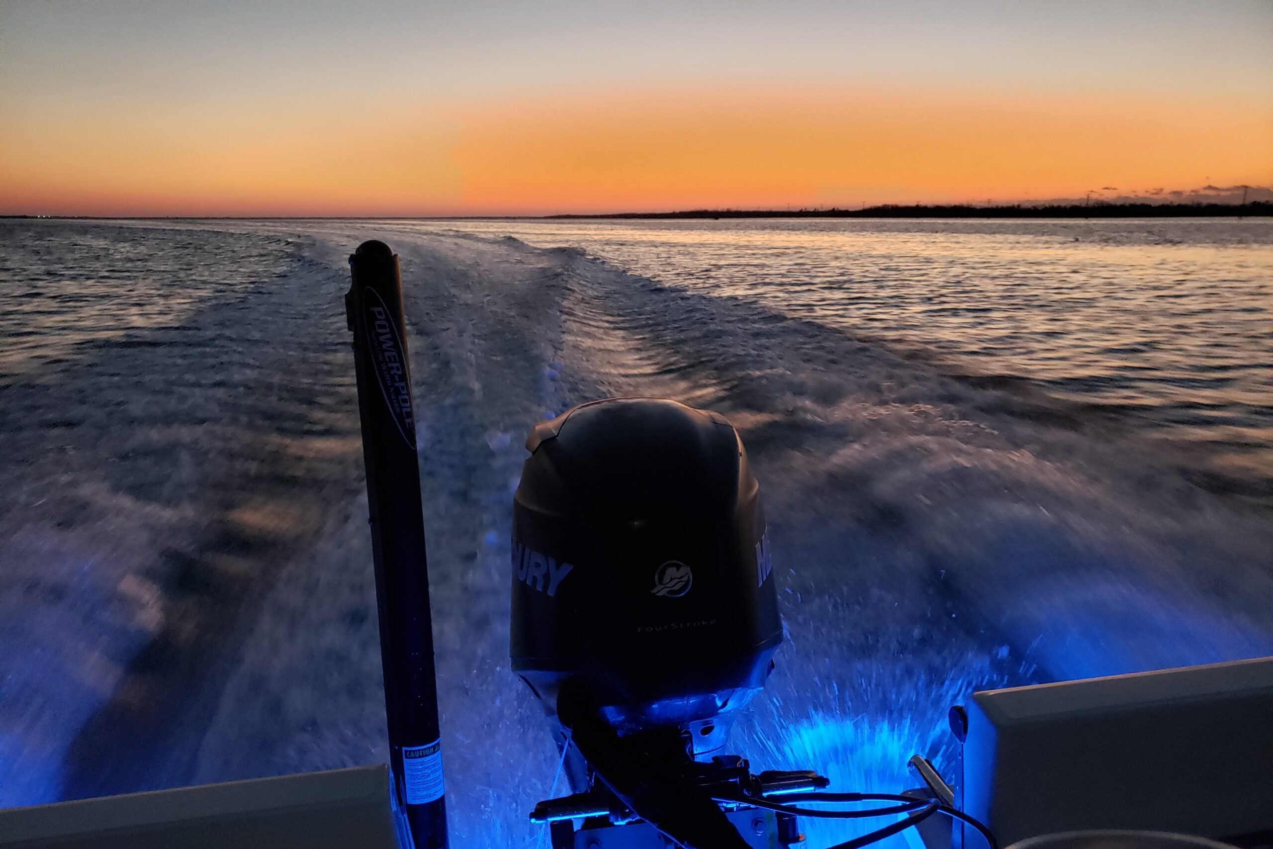 Night Cruise Glow – After-Hours on Piano Piano Mercury engine on a boat glowing with blue underwater lights during a nighttime cruise through the Gulf of Mexico at sunset.