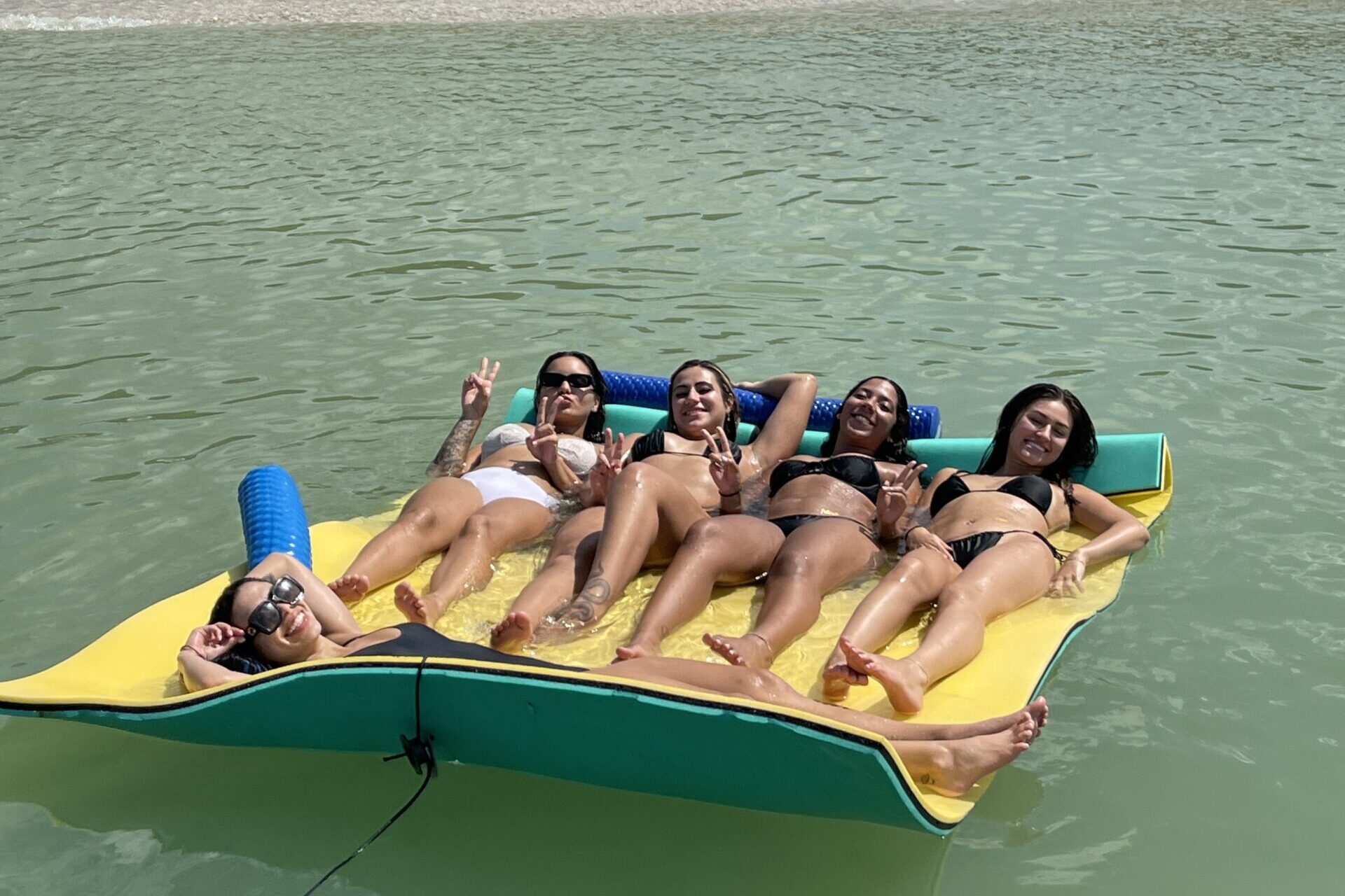 Salty Send-Off – Bachelorette Bash on the Water Five women in swimsuits smiling and throwing peace signs while lounging together on a floating mat near a beach in Naples, Florida.