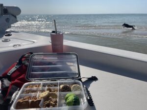 Snacks, Sun, and Salty Tails Waterproof snack tackle box on a boat with a tumbler and a dog running along the beach in the background