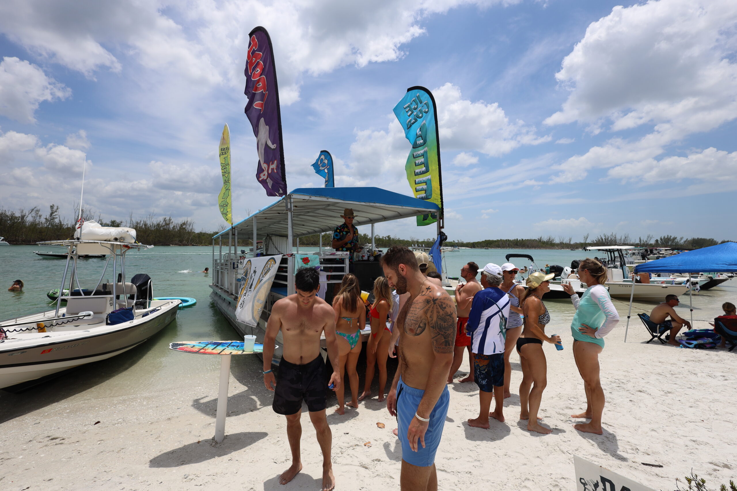 Keewaydin Party Scene – Where Sand Meets Sound Crowd of people gathered around a floating bar at Keewaydin Island with boats anchored nearby on a sunny day in Naples, Florida.
