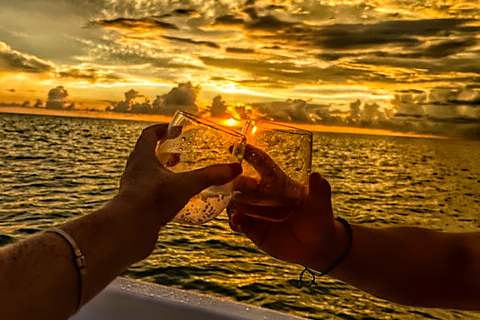 Golden Toast – Romance at Sea Two hands clinking cocktail glasses at sunset on a boat with golden skies and calm ocean water in the background.