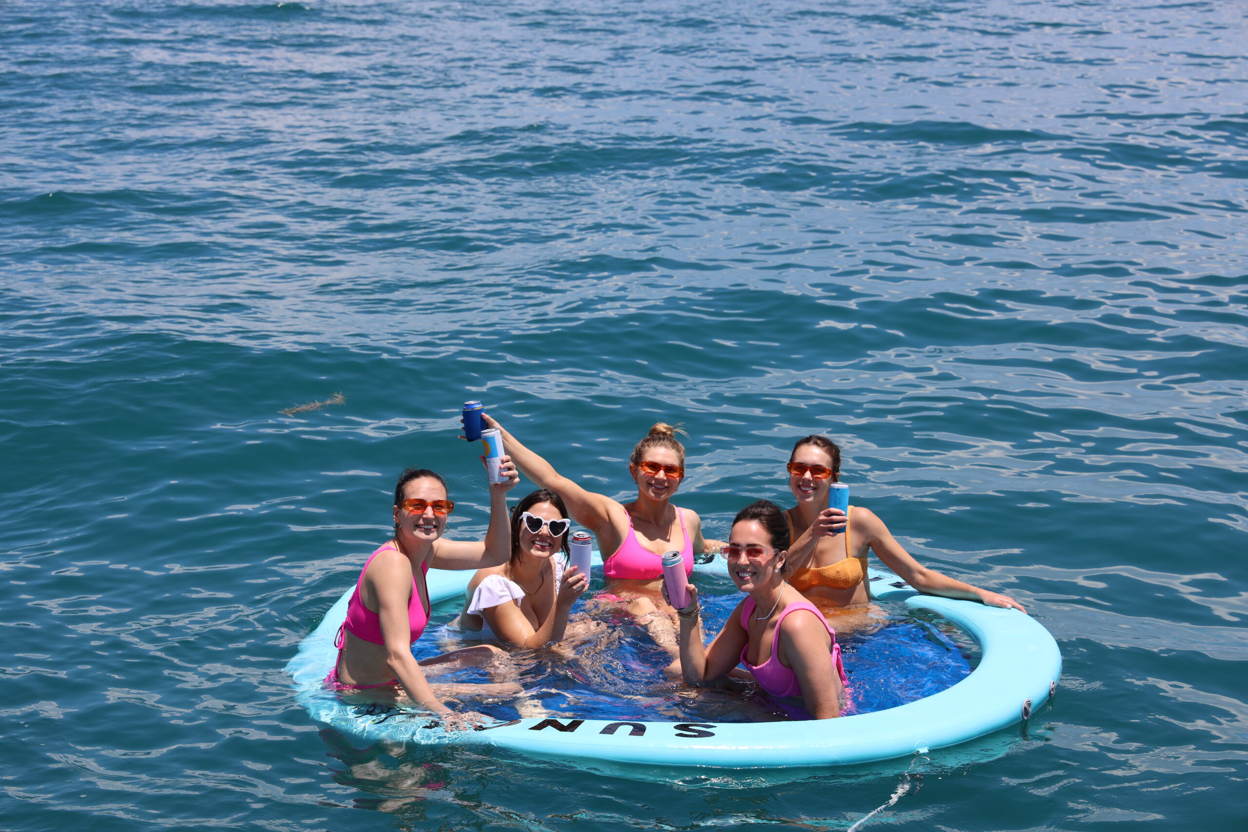 Gulf Float with the Crew – Where the Vibe Drifts with You Group of women smiling and holding drinks while floating together on a large inflatable in the Gulf of Mexico on a sunny day.