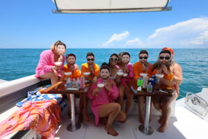 Group of women on a bachelorette boat party in Naples, FL, wearing pink and orange outfits and holding fun face masks while toasting drinks.